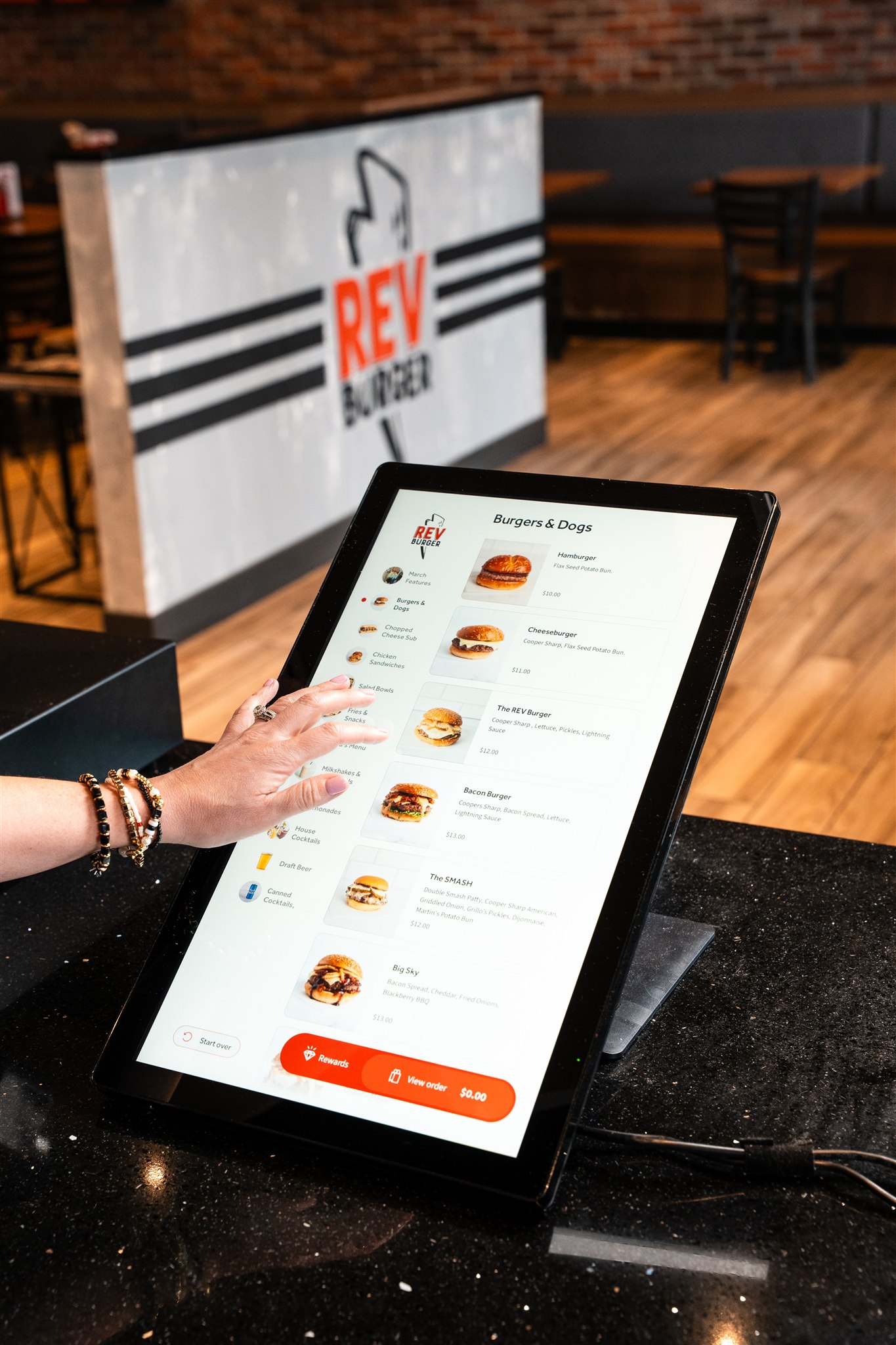 Customer using the self-order touchscreen kiosk at Rev Burger, browsing the Burgers & Dogs menu