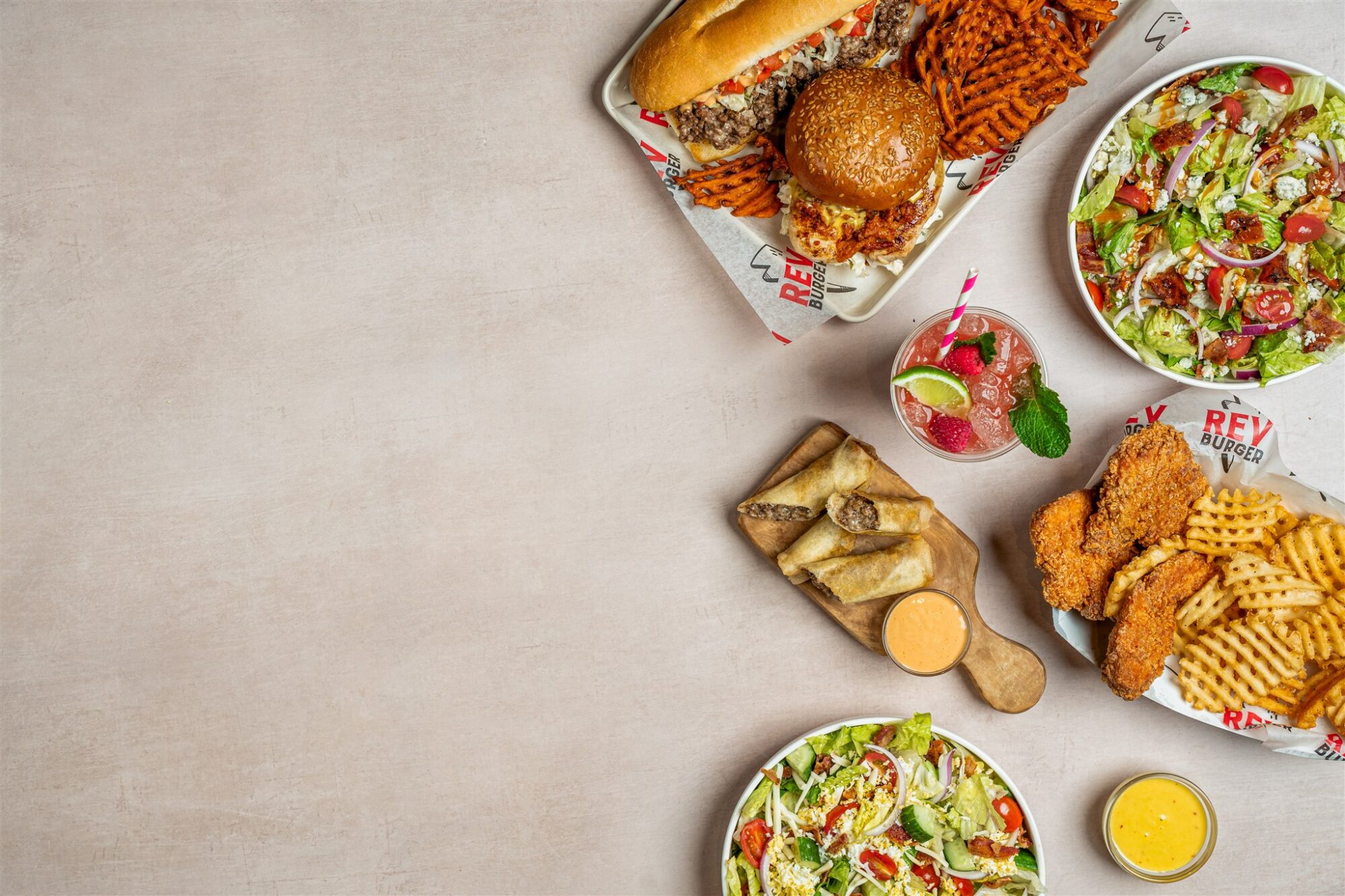 Flat lay of a Rev Burger meal spread including burgers, waffle fries, salads, chicken tenders, Cheeseburger Spring Rolls, and a raspberry rickey