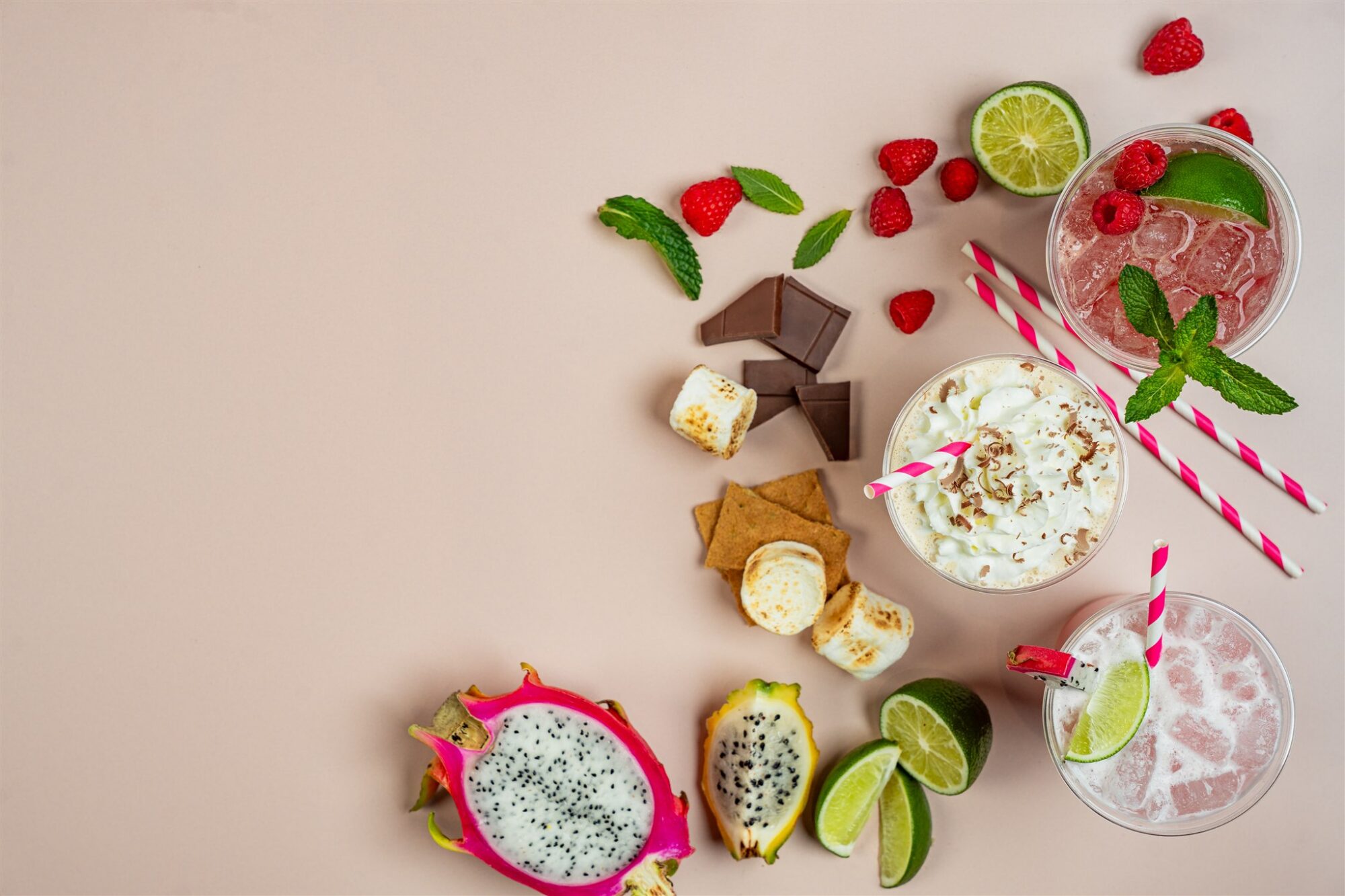 Flat lay of specialty drinks with fresh raspberries, lime, dragon fruit, and s'mores ingredients on a pink background